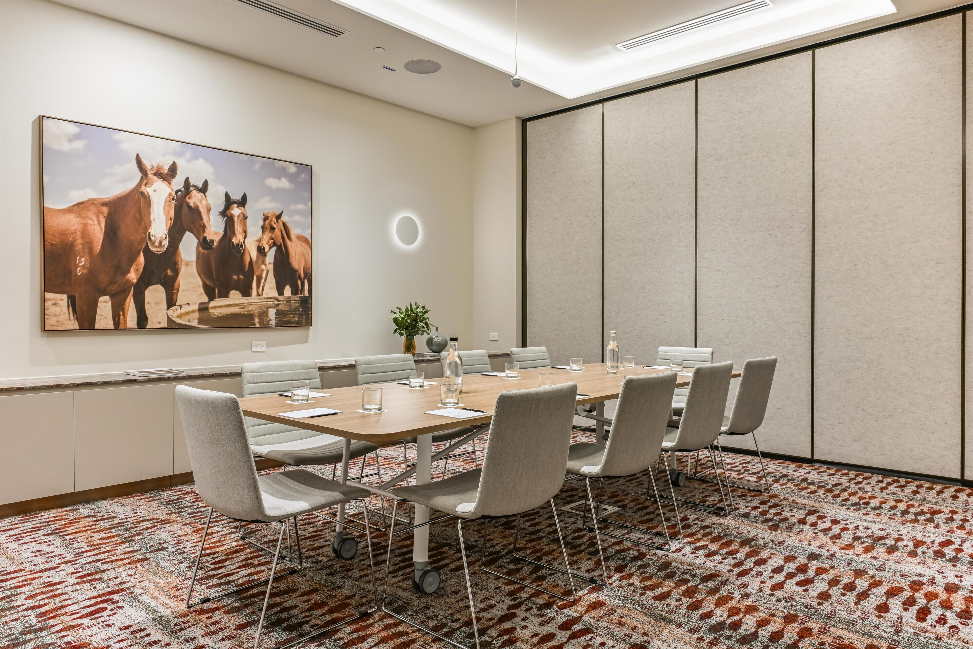 A conference room with a long table, chairs, a large painting of horses, and a patterned carpet.
