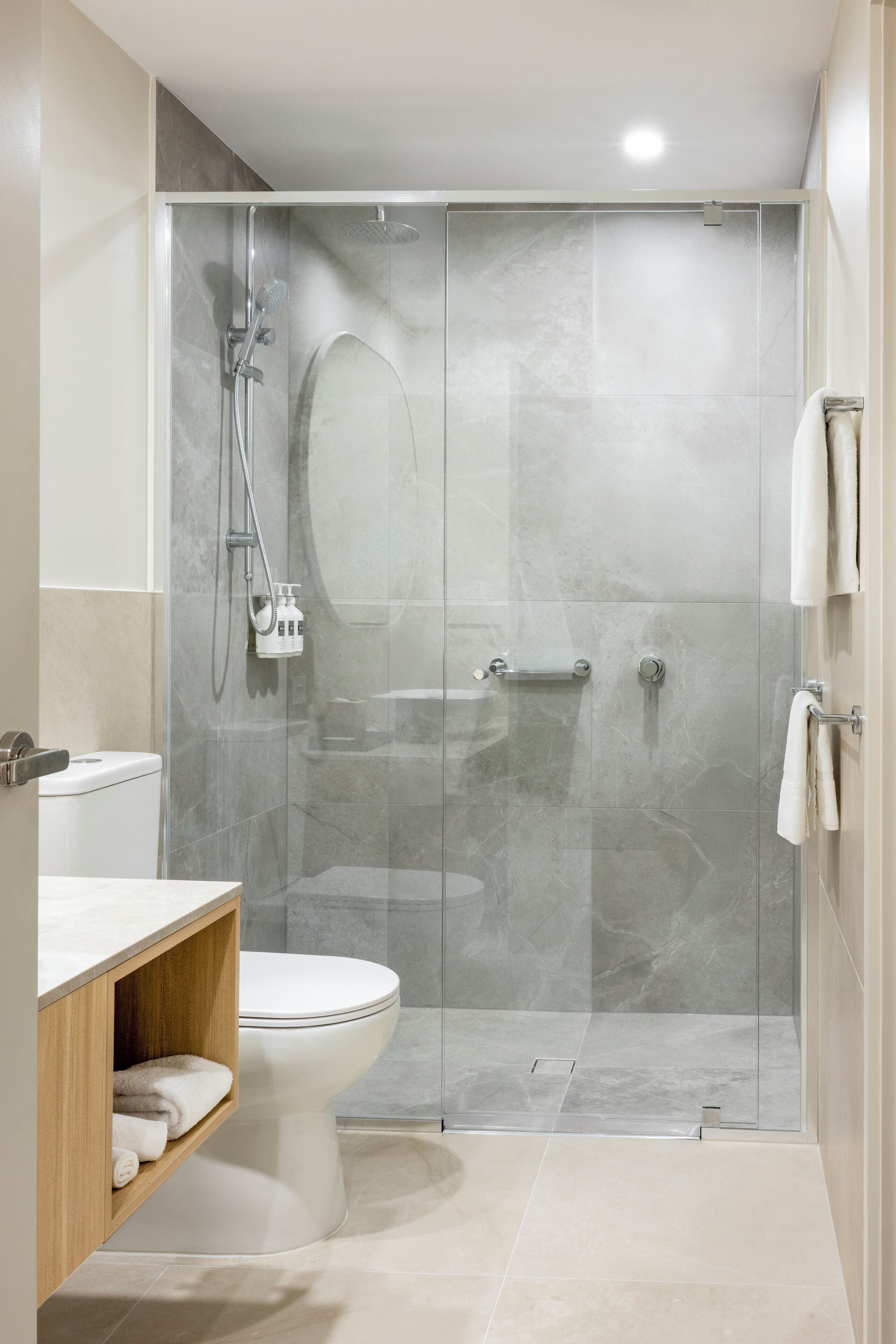 A modern bathroom with a glass-enclosed shower, a white toilet, and a wooden vanity with a shelf.
