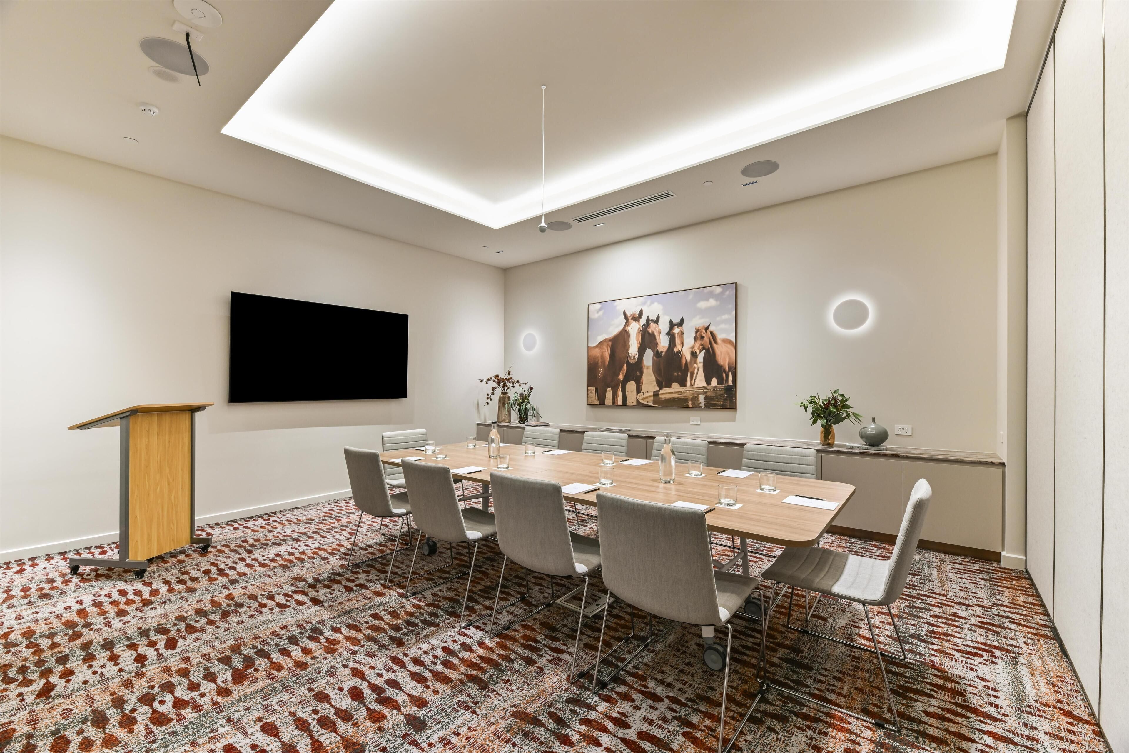 A conference room with a long table, chairs, a podium, a flat-screen TV, and a painting on the wall.