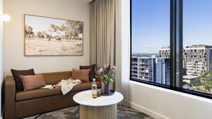 A modern hotel room with a large window, a sofa with cushions, a small round table with a vase and bottle, and a painting on the wall. - Adina Chermside Brisbane (Brisbane)
