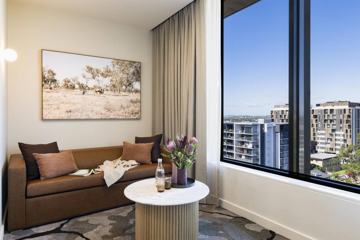 A modern hotel room with a large window, a sofa with cushions, a small round table with a vase and bottle, and a painting on the wall.
