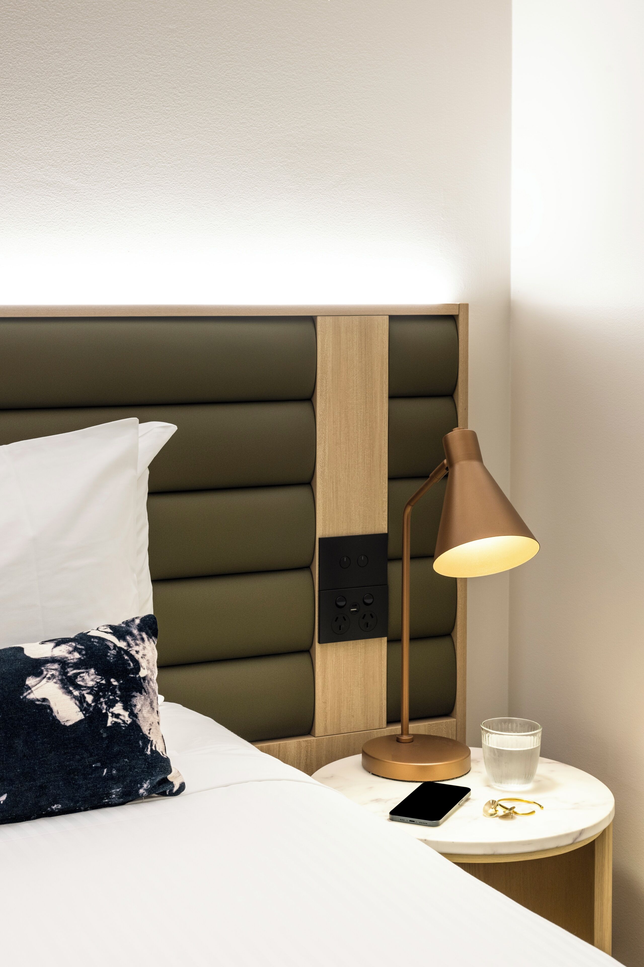 A neatly made bed with a pillow, a bedside lamp, a glass of water, and a smartphone on a bedside table.