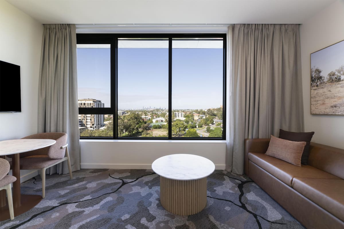 A hotel room with a large window, a sofa, a round table, and a TV.