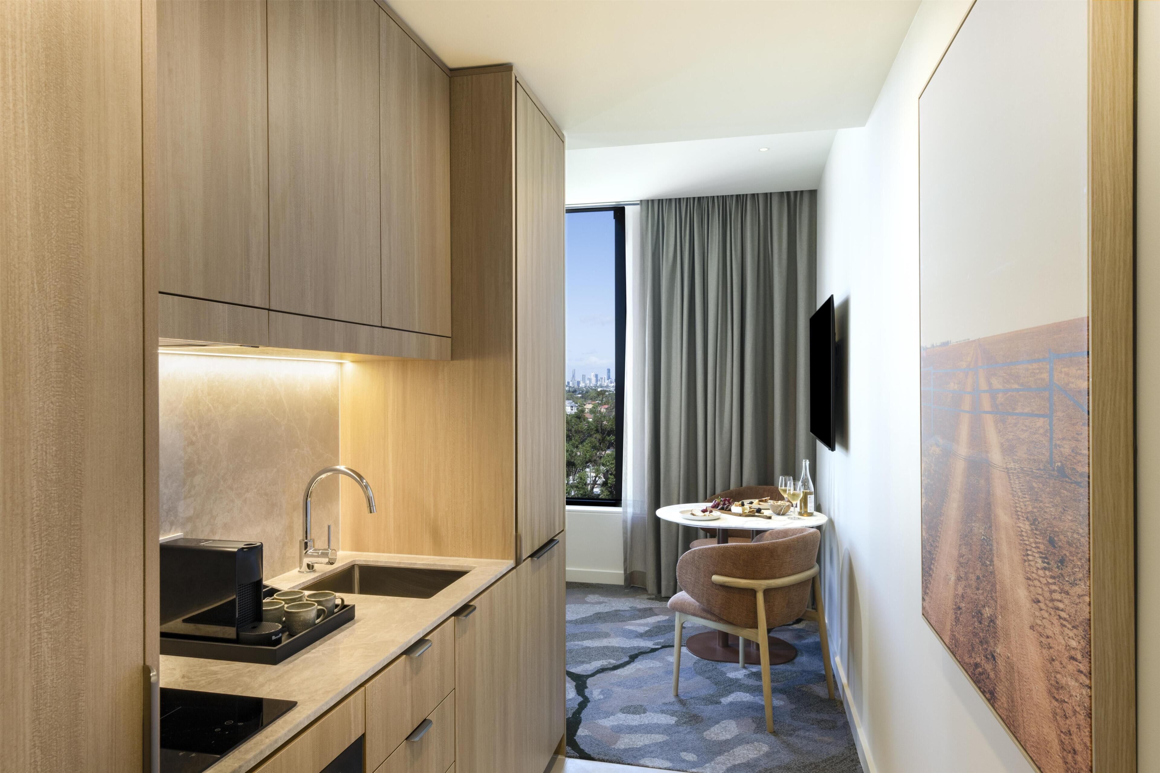 A compact hotel room with a kitchenette, a dining area with a table and chairs, a large window with curtains, and a mounted television.