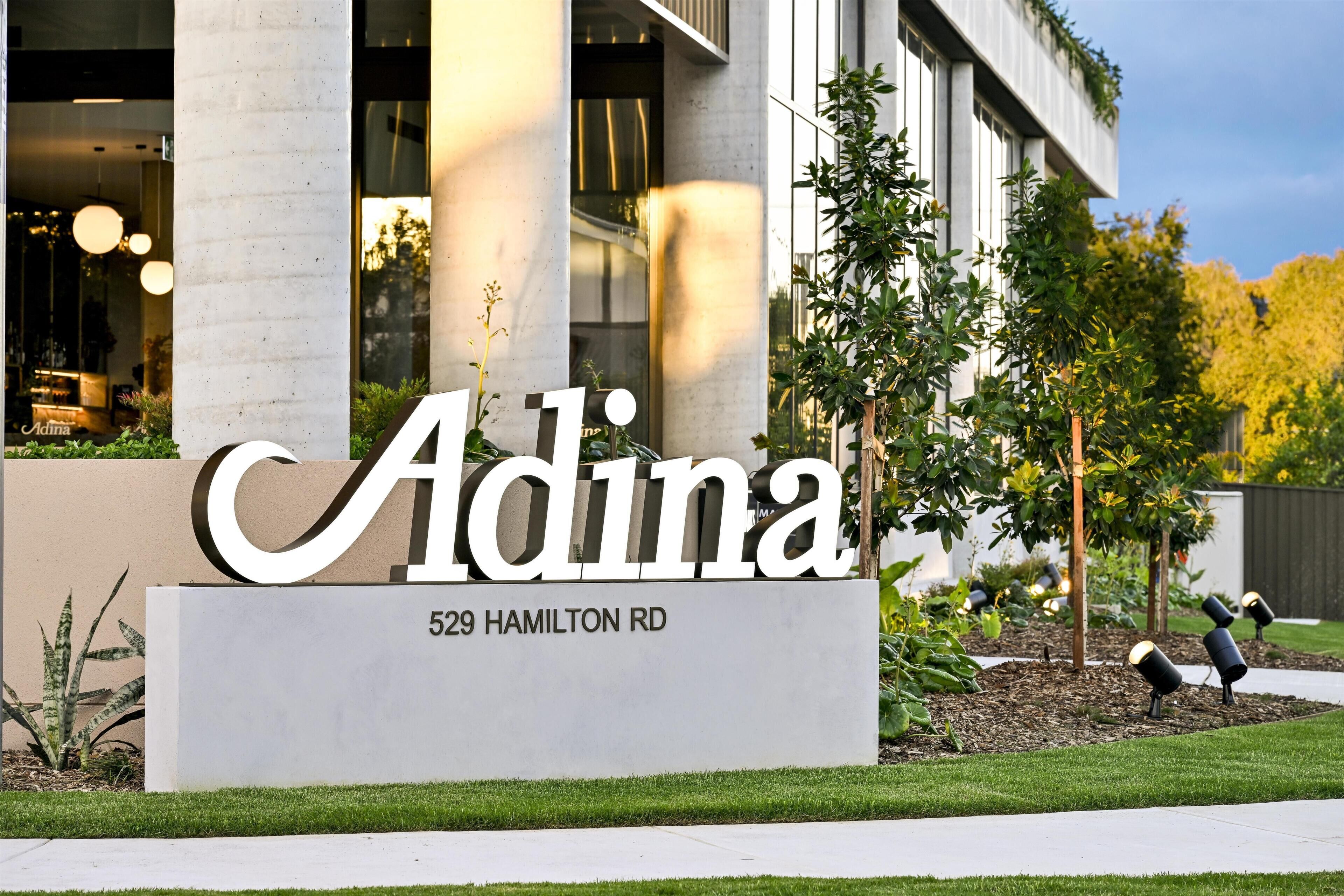 En bygning med et stort skilt, der viser 'Adina' og '529 Hamilton Rd', foran en moderne bygning med store vinduer og søjler.