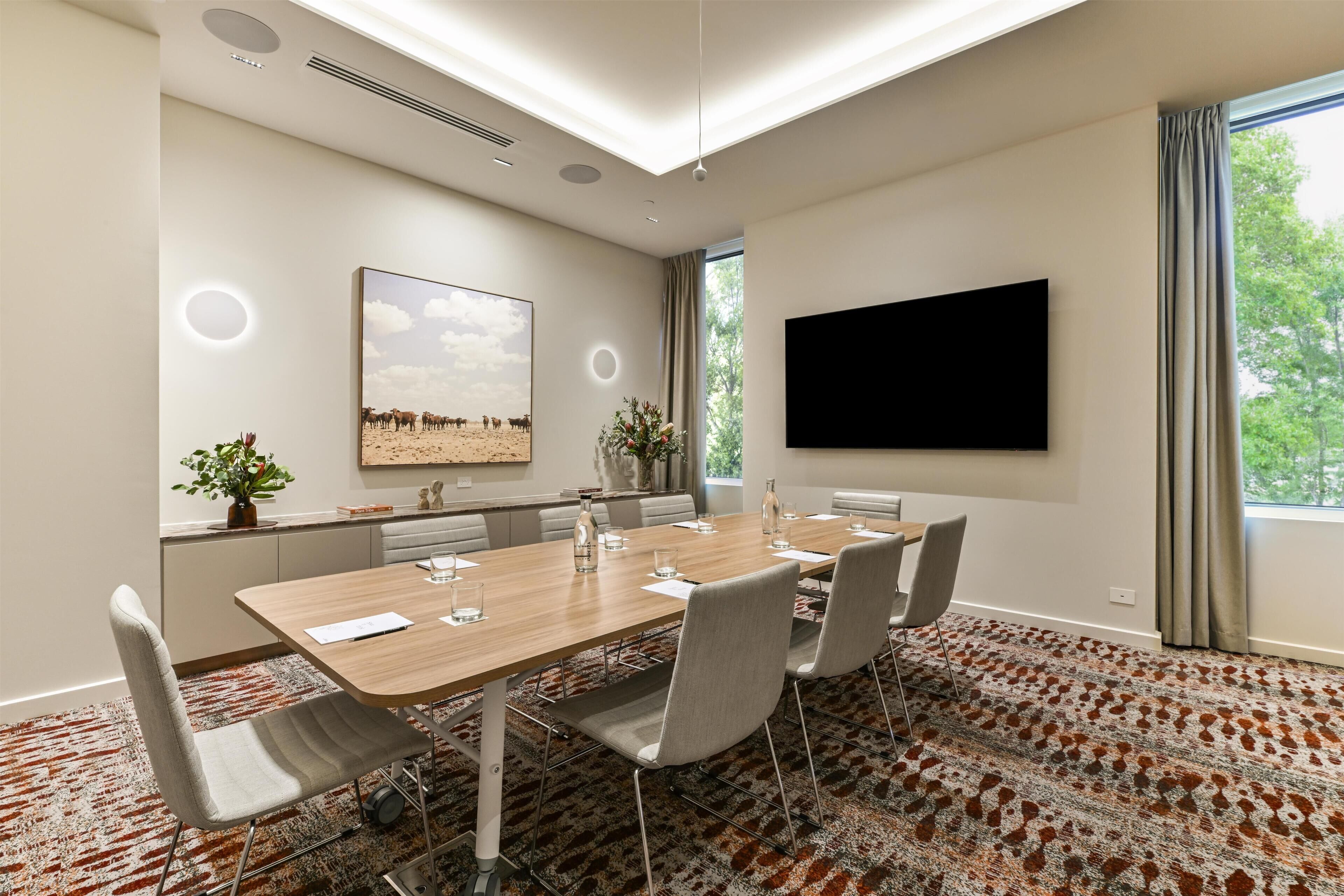A conference room with a long table, chairs, a flat-screen TV, a painting, and a window with curtains.