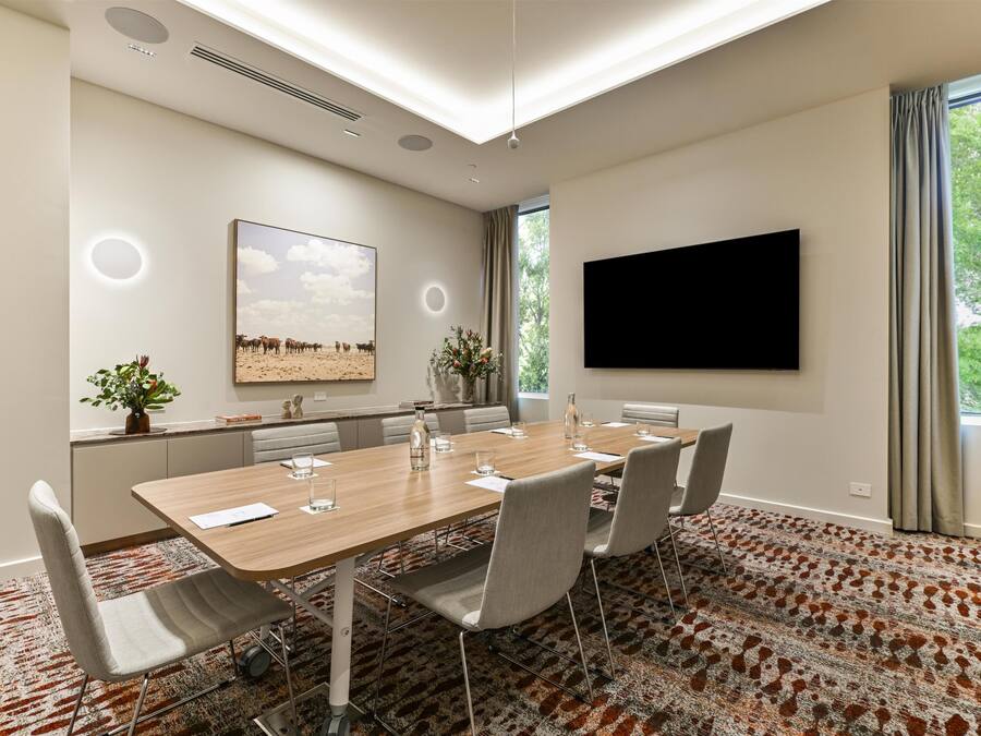 Un salón de conferencias con una larga mesa, sillas, un televisor de pantalla plana, un cuadro y una ventana con cortinas.