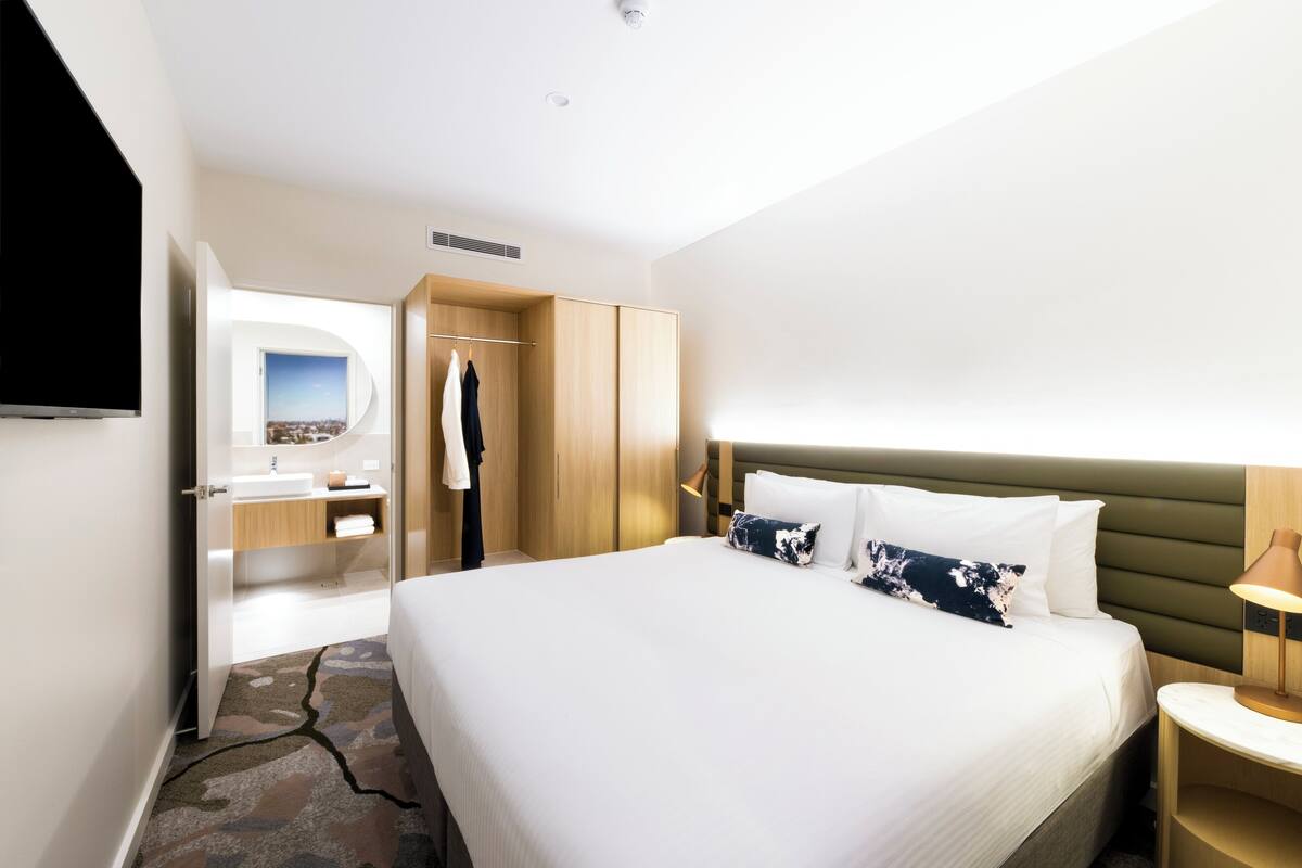 A modern hotel room with a large bed, a television, a bathroom with a sink and mirror, and a view of the city.