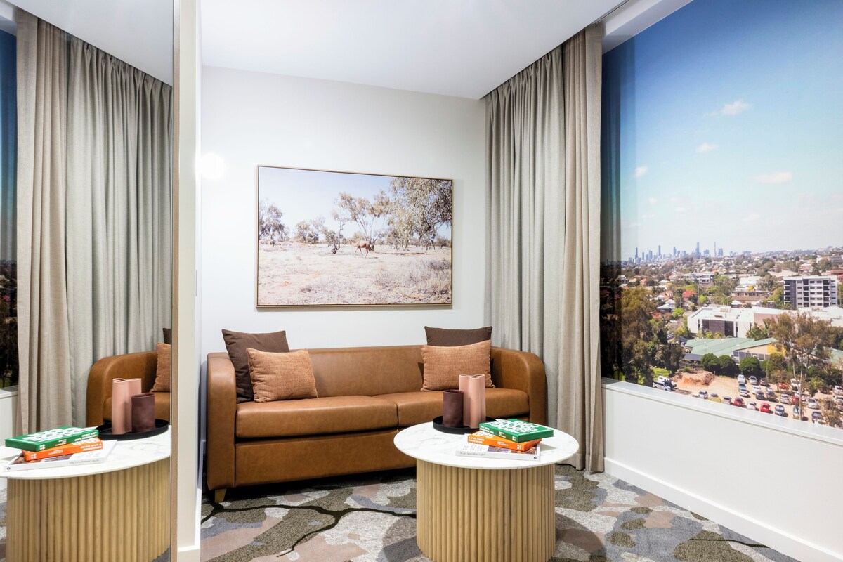 A modern hotel room with a brown leather sofa, a round coffee table, and a large window with a cityscape view.