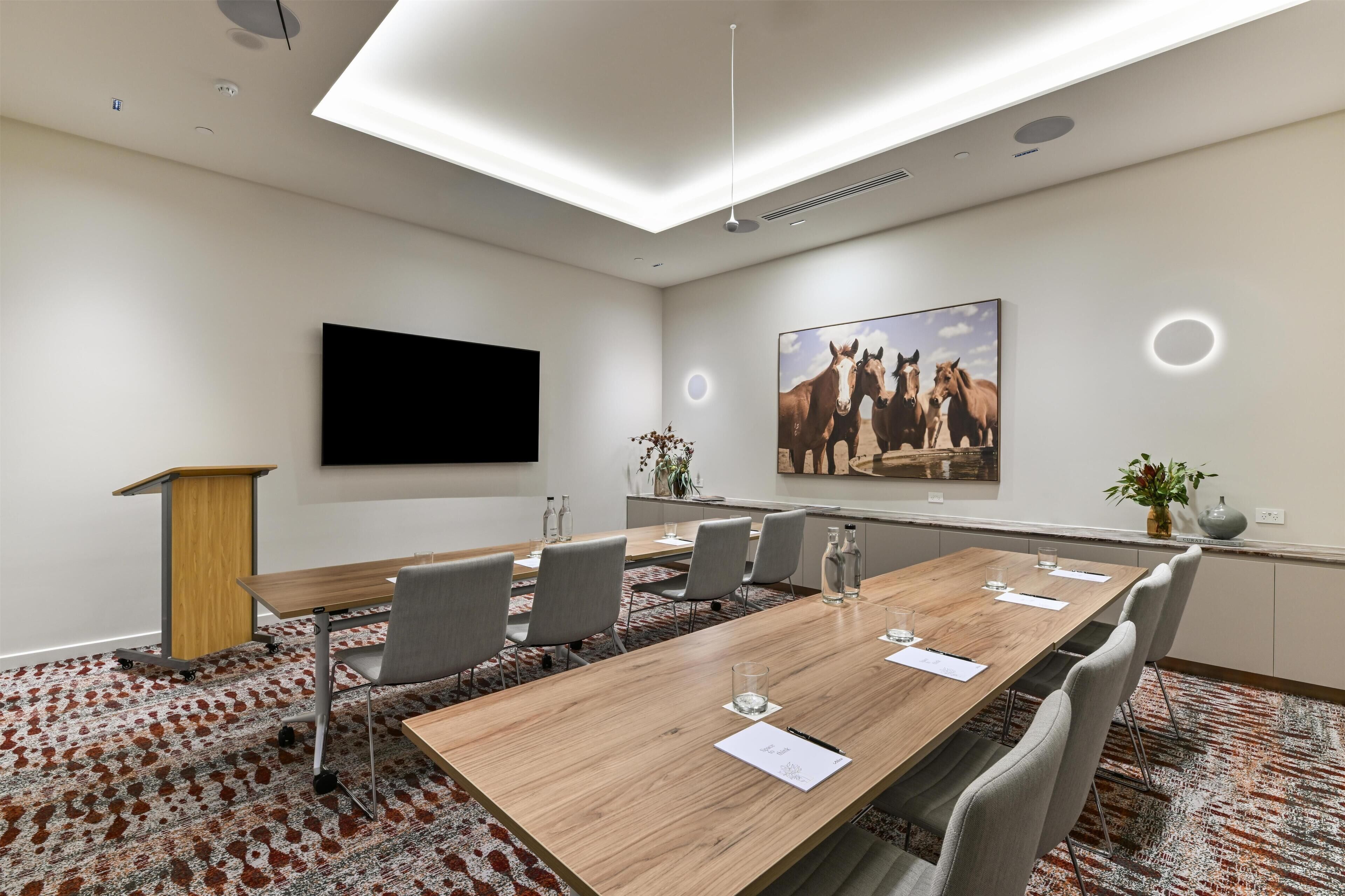 Une salle de conférence avec une longue table, des chaises, un pupitre, un téléviseur à écran plat et un tableau représentant des chevaux.
