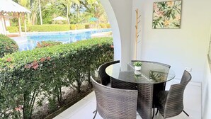 Ein Balkon mit Glastisch und Korbstühlen, der auf einen Swimmingpool und üppiges Grün blickt.