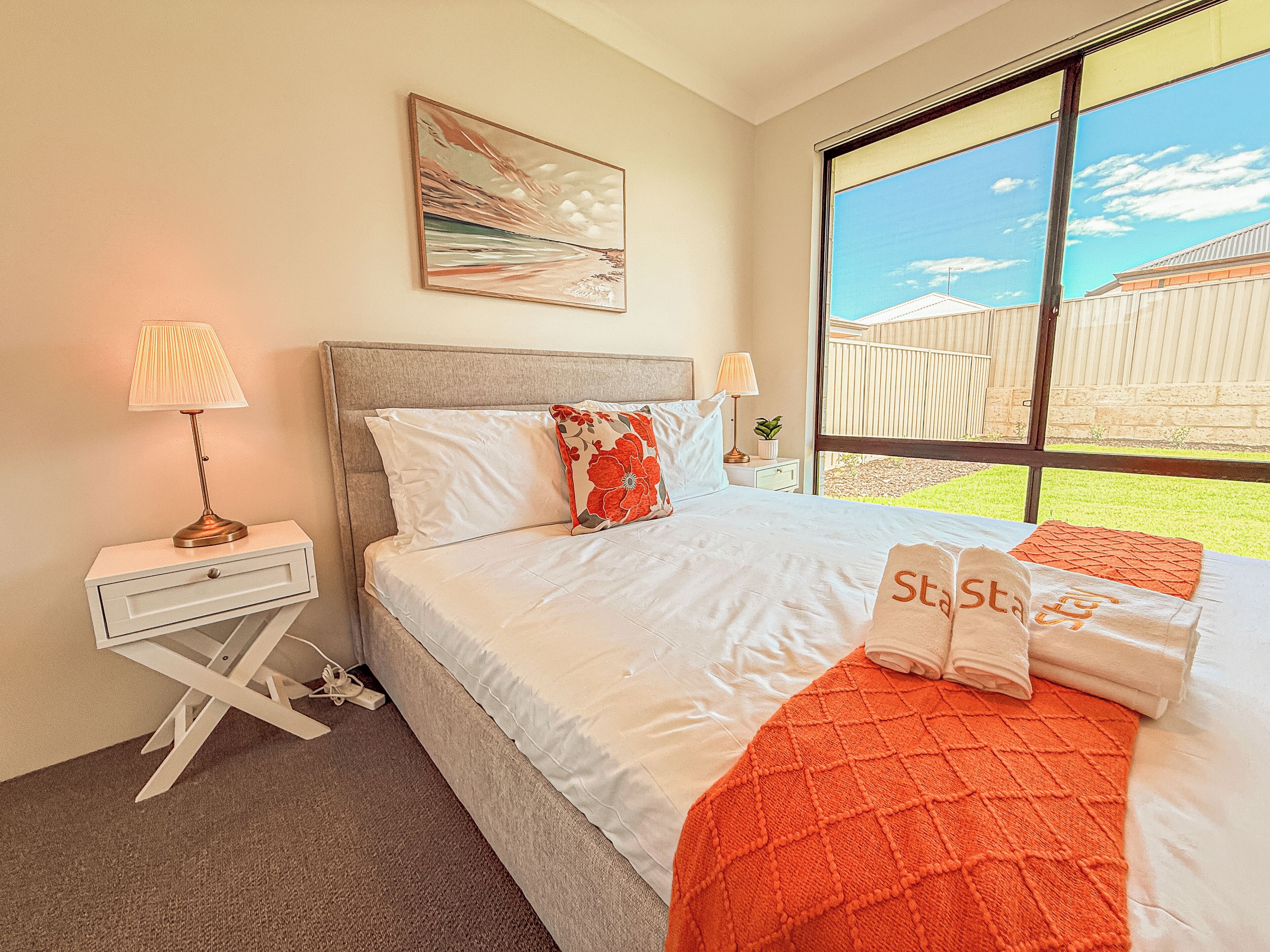 Un dormitorio con cama, mesita de noche, lámpara, ventana con vistas al exterior y un cuadro en la pared.