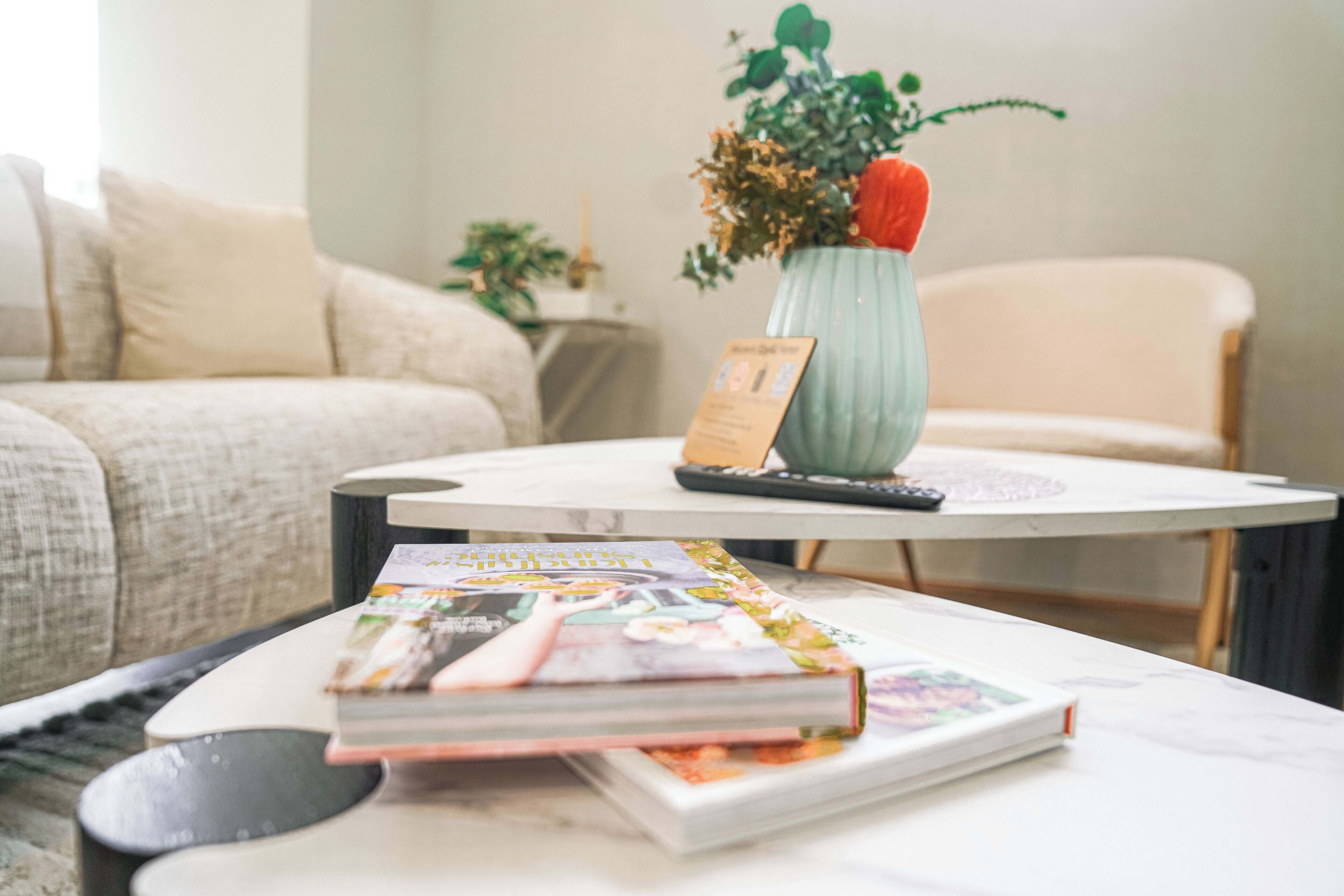 A living room with a sofa, a coffee table with magazines, a vase with flowers, and a remote control.