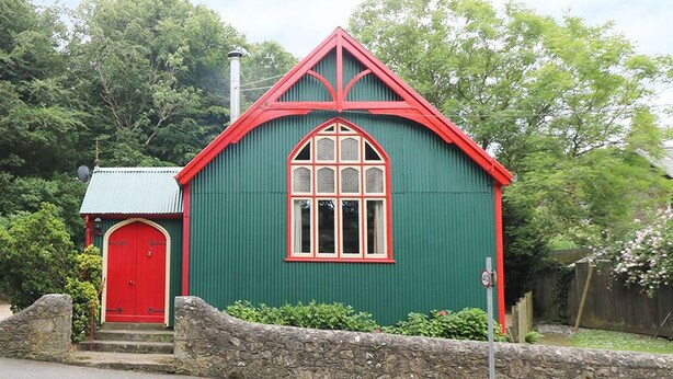 A green building with a red door and window frames, a stone wall in front, and a small attached structure with a green roof. - The Mission (Ventnor)