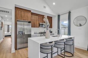 A modern kitchen with a central island, bar stools, a refrigerator, and wooden cabinets. - Mega Market 6BR Home in Houston Groups Sleeps 12+ (Houston)