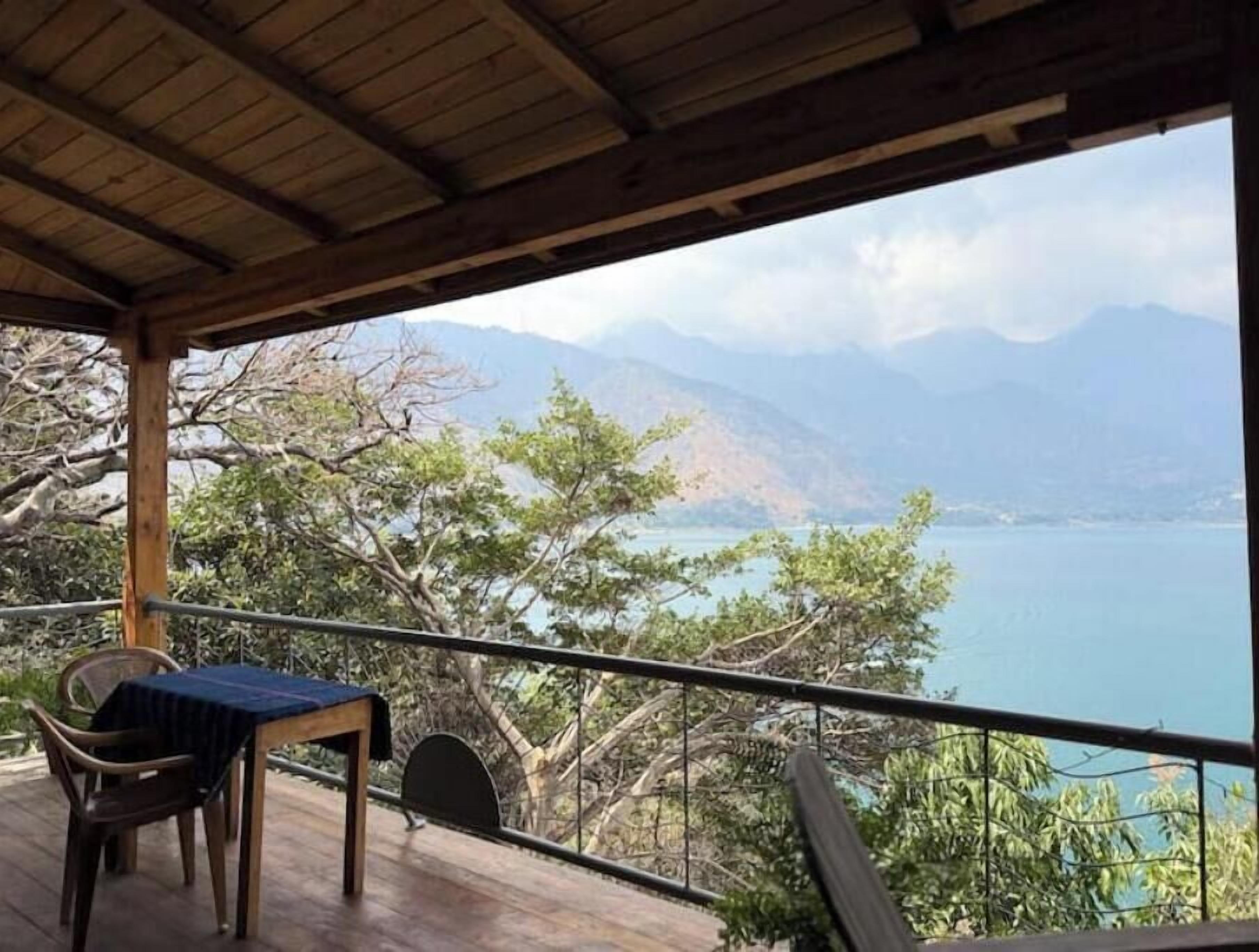 Ein überdachter Außenbereich mit Tisch und Stühlen, mit Blick auf ein Gewässer und Berge.