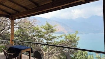 Ein überdachter Außenbereich mit Tisch und Stühlen, mit Blick auf ein Gewässer und Berge.