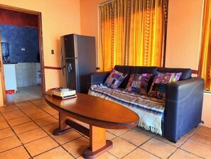 Un salon avec un canapé, une table basse, un réfrigérateur et un coin cuisine à l’arrière-plan.