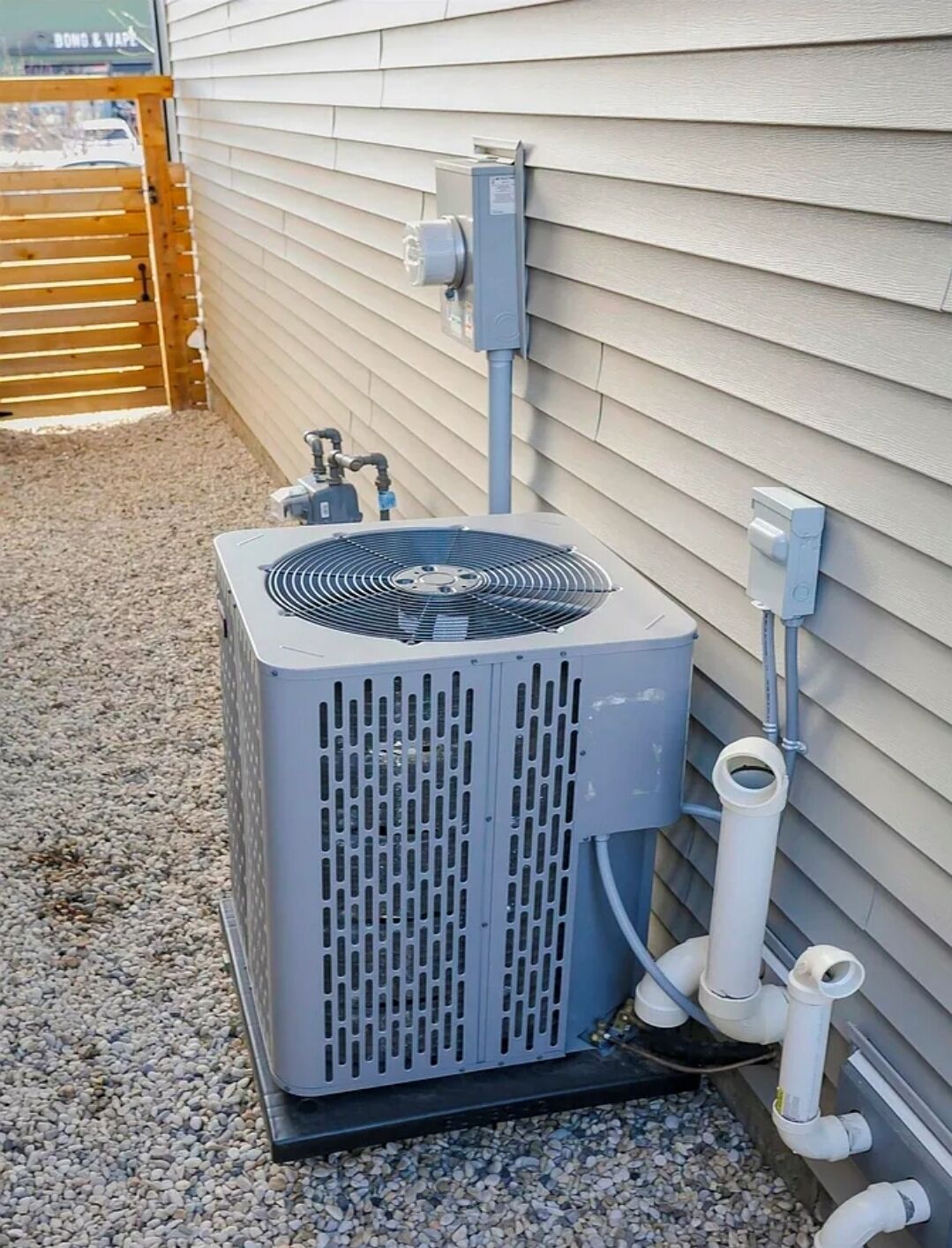 An outdoor air conditioning unit mounted on a house's exterior wall, connected to a utility box and pipes.