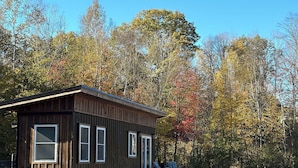 A wooden cabin with a deck, surrounded by a grassy field and autumn-colored trees.