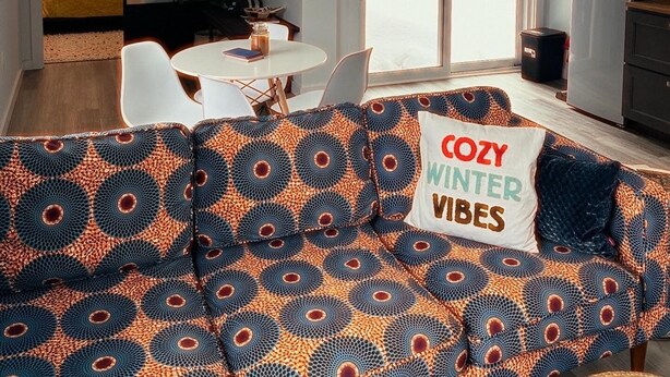 A cozy living room with a patterned sofa, a ceiling fan, and a large window with a view of trees.