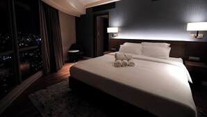 A hotel room with a large bed, two bedside lamps, a nightstand, a small table, and a view of the city at night. - The Hotel Residences at Aqua - Premier Suite  (Mandaluyong City)