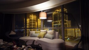 A modern living room with a sectional sofa, a coffee table with decorative items, and a large window offering a view of illuminated city buildings at night. - The Hotel Residences at Aqua - Premier Suite  (Mandaluyong City)
