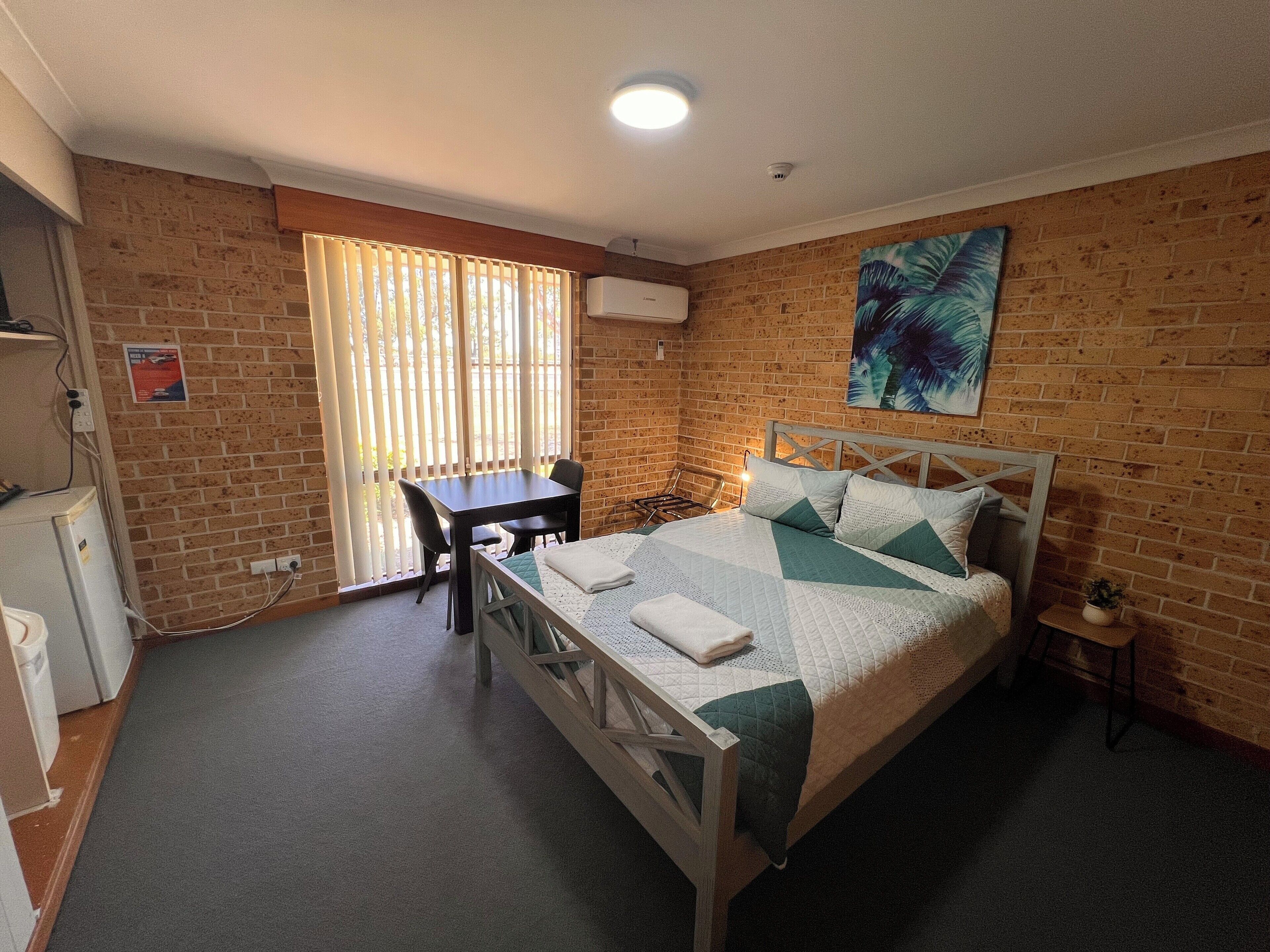 A room with a bed, a desk, and a chair. There is a window with blinds, a wall-mounted air conditioner, and a painting on the wall.