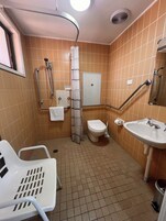 Un bagno piccolo con doccia, toilette e lavandino. Si nota una sedia bianca e un cestino della spazzatura bianco.