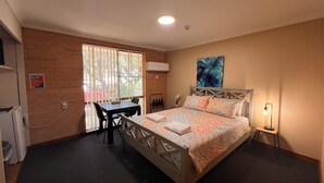 Une chambre à coucher avec un lit, un bureau et une chaise. Il y a une fenêtre avec des stores, un tableau au mur et une lampe sur la table de chevet.