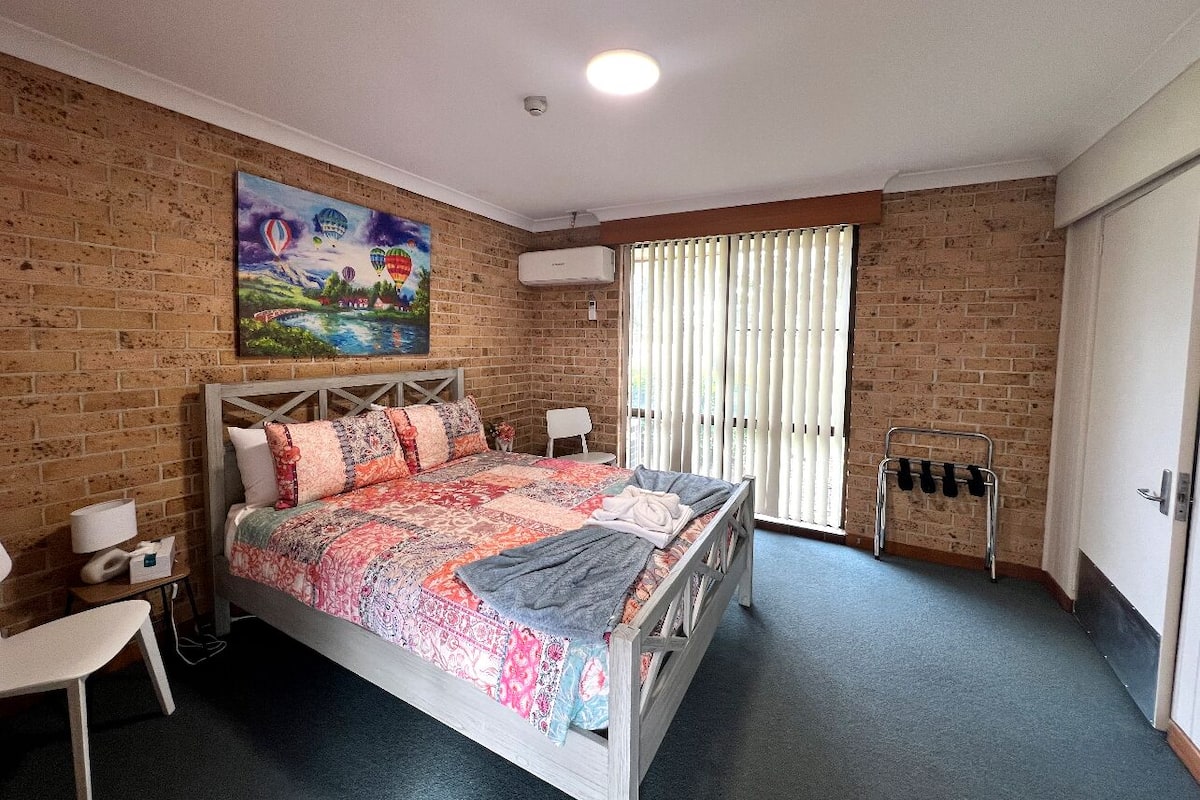 A bedroom with a brick wall, a bed with a colorful quilt, a painting above the bed, a window with blinds, and a coat rack.