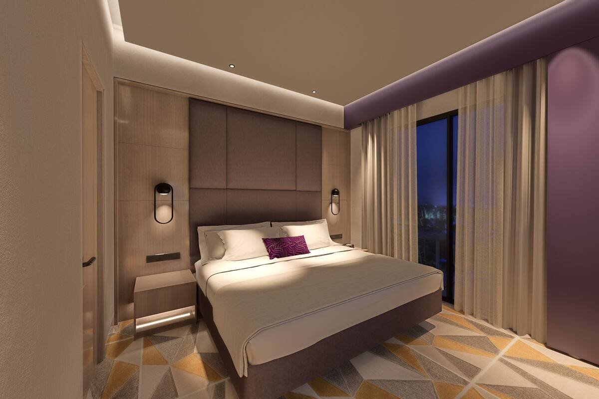 Une chambre d’hôtel moderne avec un grand lit, une table de chevet, une lampe fixée au mur et une vue sur la ville la nuit.