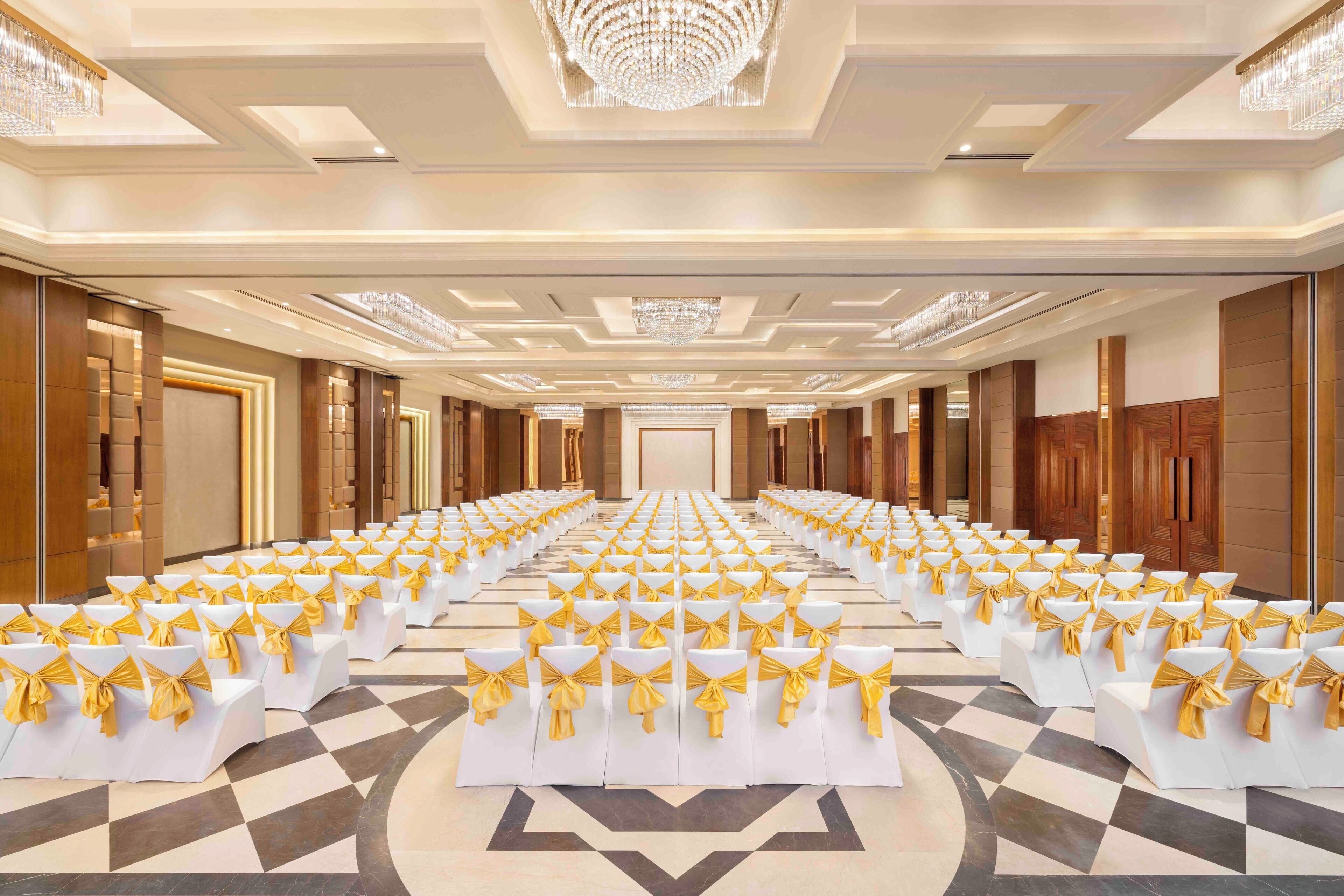 A spacious hall with rows of white chairs and yellow sashes, a chandelier, and a checkered floor pattern.