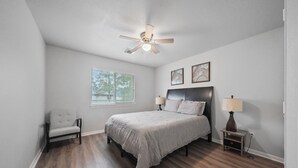 A bedroom with a bed, a chair, a nightstand, a ceiling fan, and a window. - Cozy, Comfy, Warm Relaxing Home (Conroe)