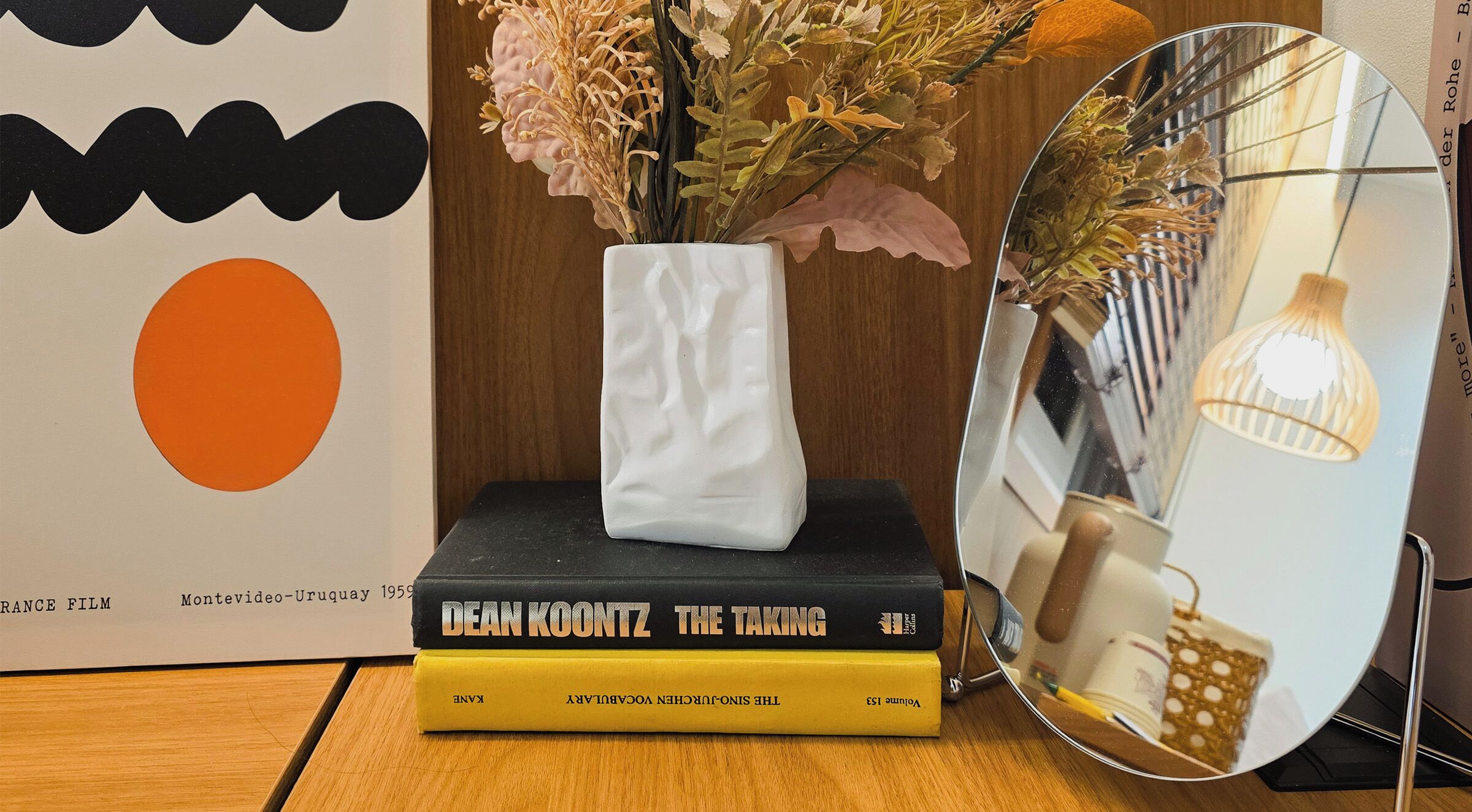 A stack of books titled 'The Taking' by Dean Koontz, placed on a wooden surface with a white vase containing dried flowers.