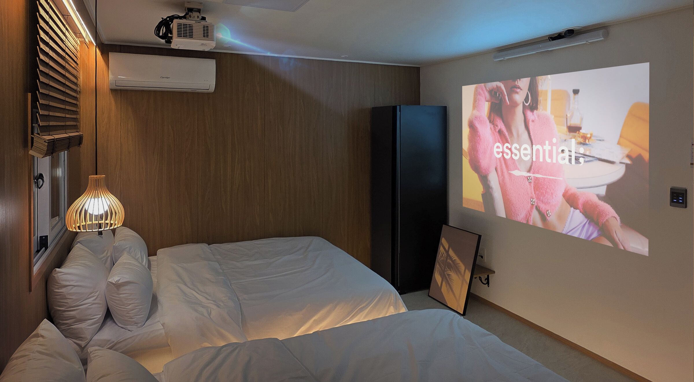 Une chambre d’hôtel équipée d’un lit, d’un écran de projection affichant une image, d’un projecteur fixé au plafond et d’un climatiseur mural.