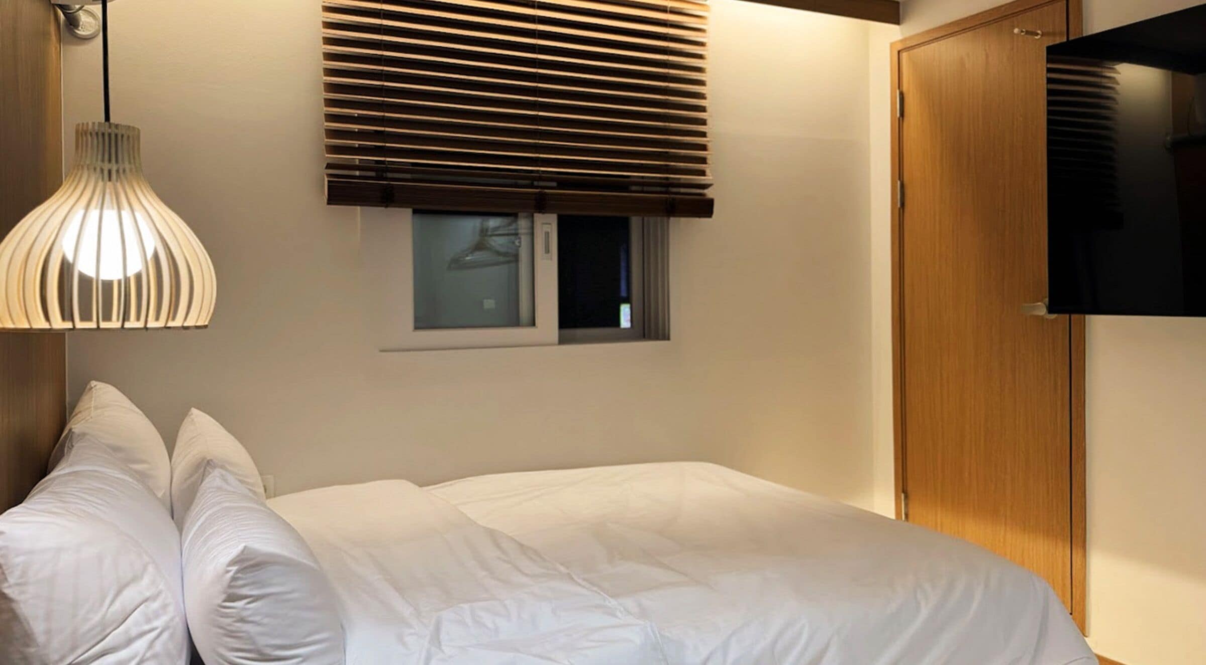 A bedroom with a bed, a window with blinds, a wall-mounted TV, and a pendant light.