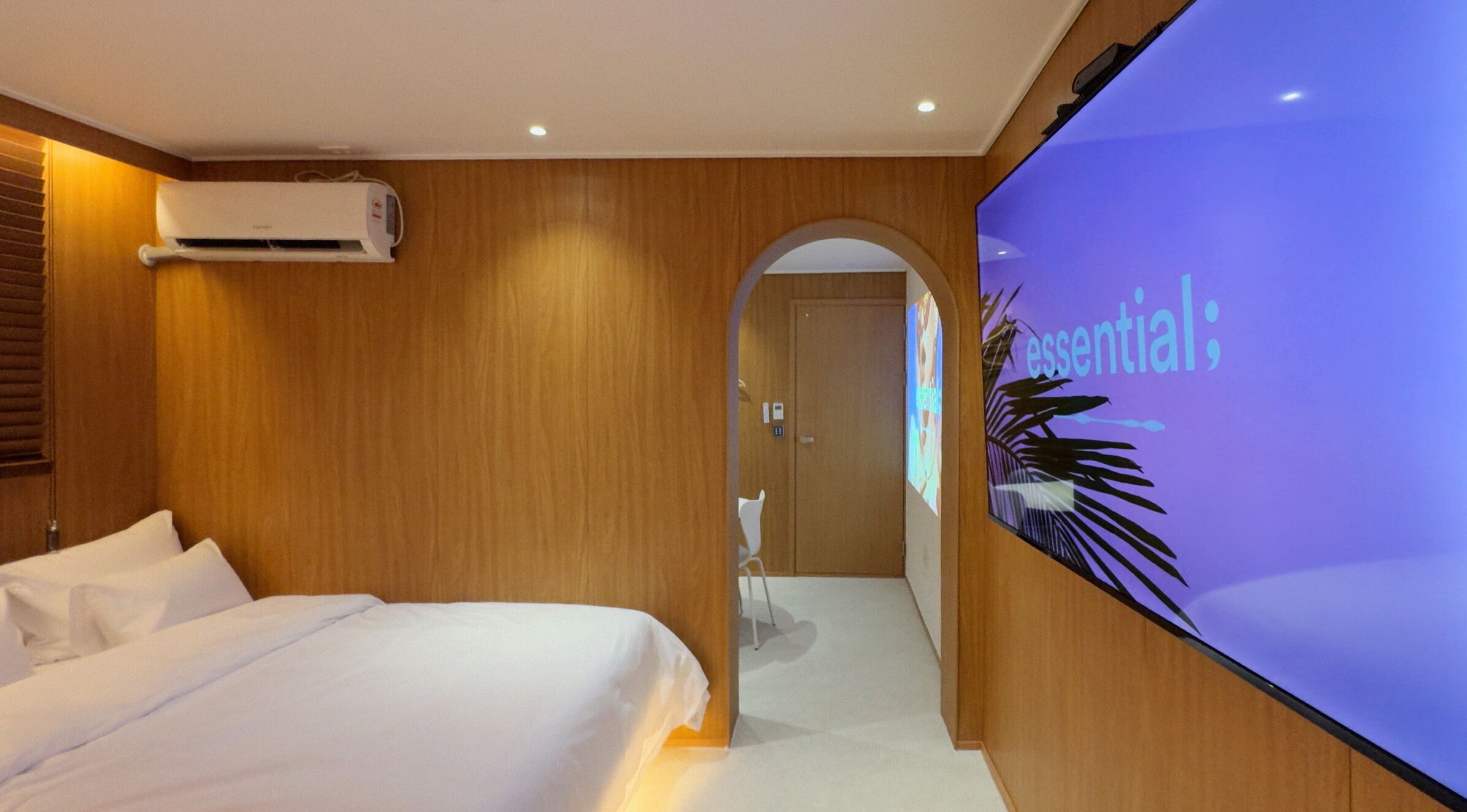 Une chambre d’hôtel avec un lit, un appareil de climatisation, un écran de télévision affichant le mot « essentiel », et un mur en bois.