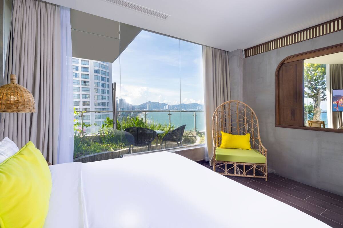 Une chambre d’hôtel avec un grand lit, un fauteuil en osier avec un coussin jaune, une télévision et une vue sur le paysage urbain et les montagnes, visible à travers de grandes fenêtres.