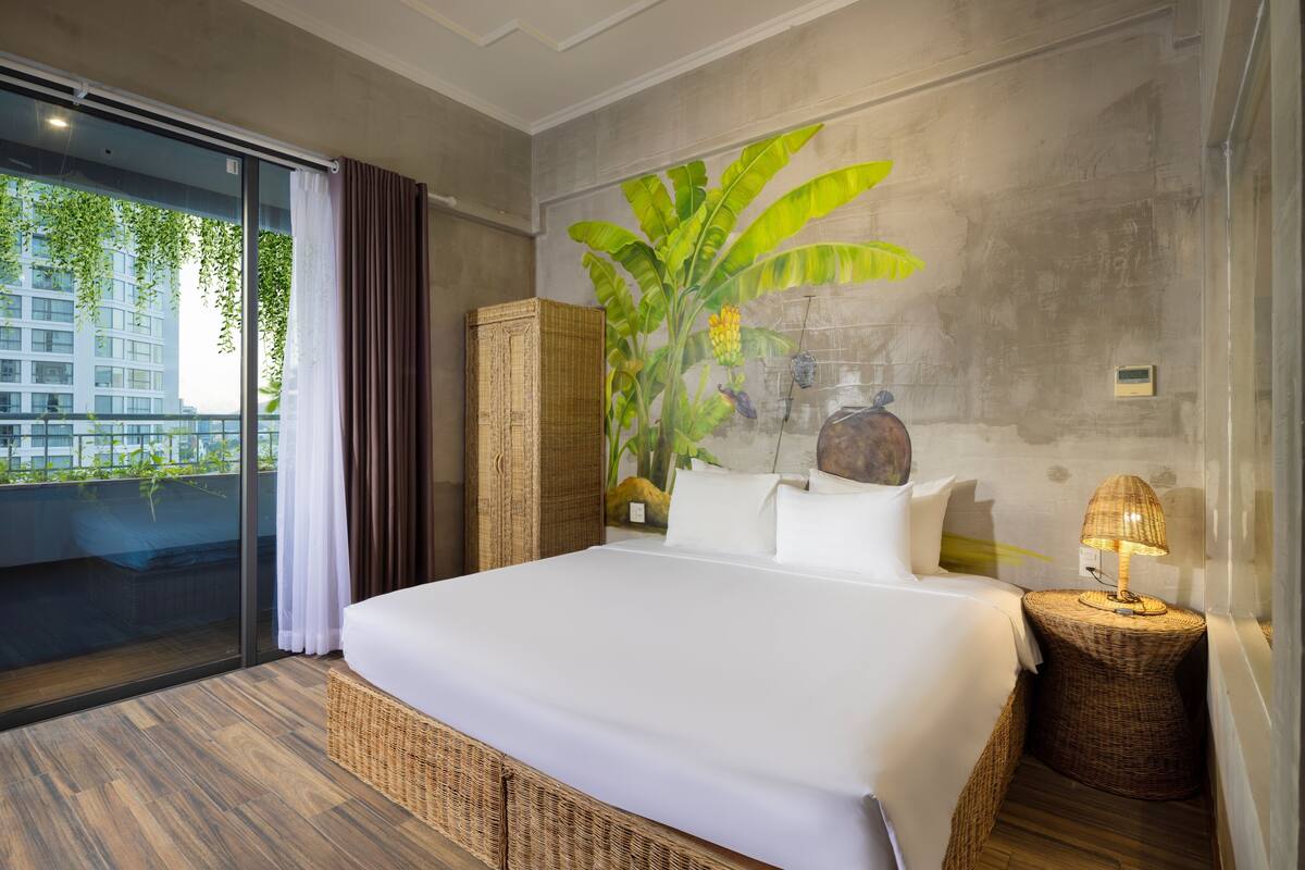 Une chambre d'hôtel moderne avec un grand lit, des meubles en osier, une plante en pot et une vue sur les bâtiments par la fenêtre.