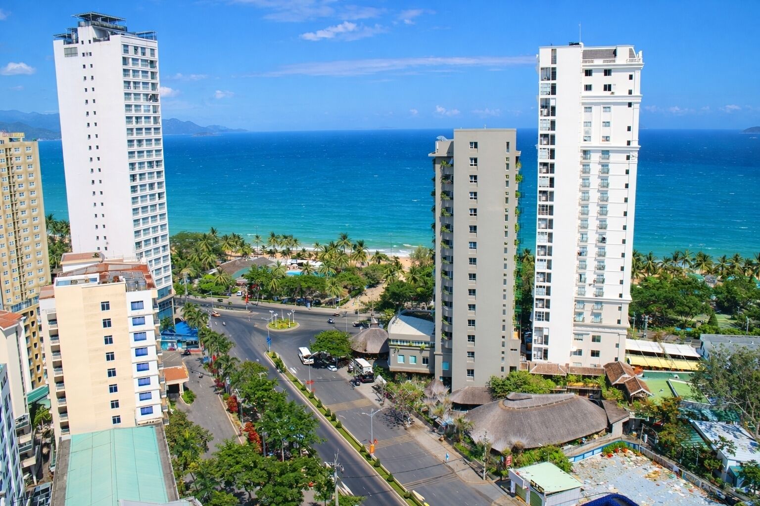 A coastal cityscape with high-rise buildings, a central road, and a beach with palm trees.