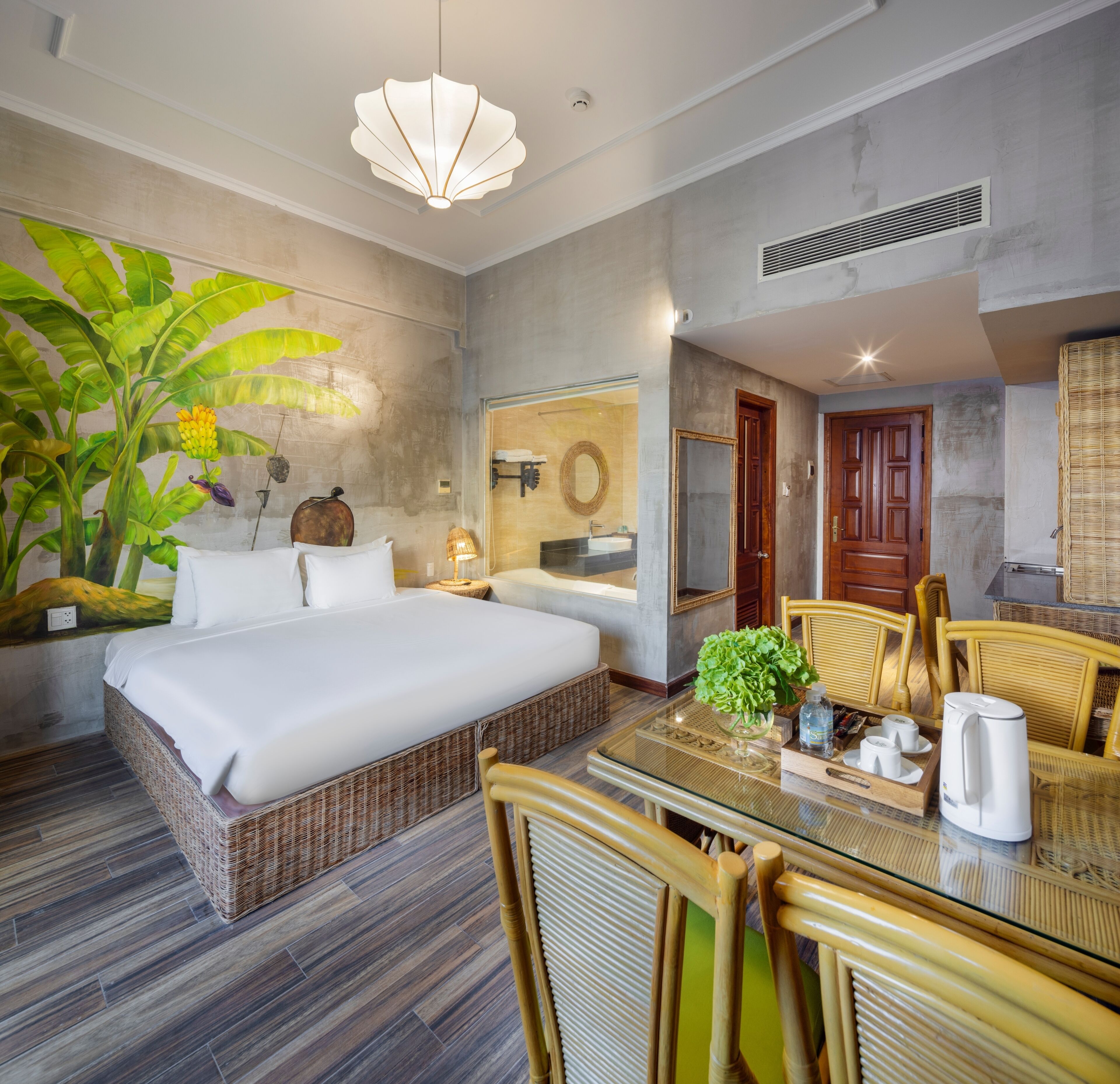 A hotel room with a large bed, a dining table with chairs, a plant, and a bathroom visible through a glass partition.