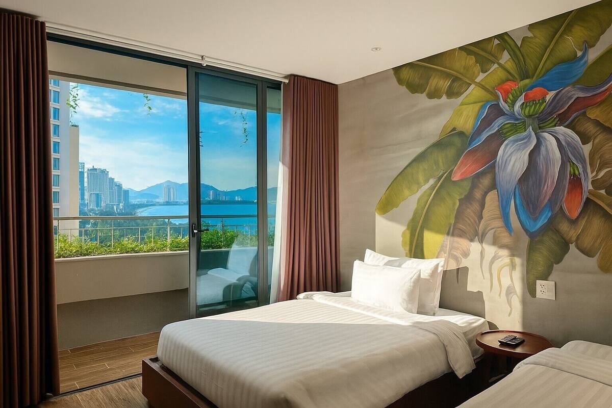 Une chambre d’hôtel avec un grand lit, une vue sur le paysage urbain à travers une porte-fenêtre coulissante et une fresque représentant une fleur sur le mur.