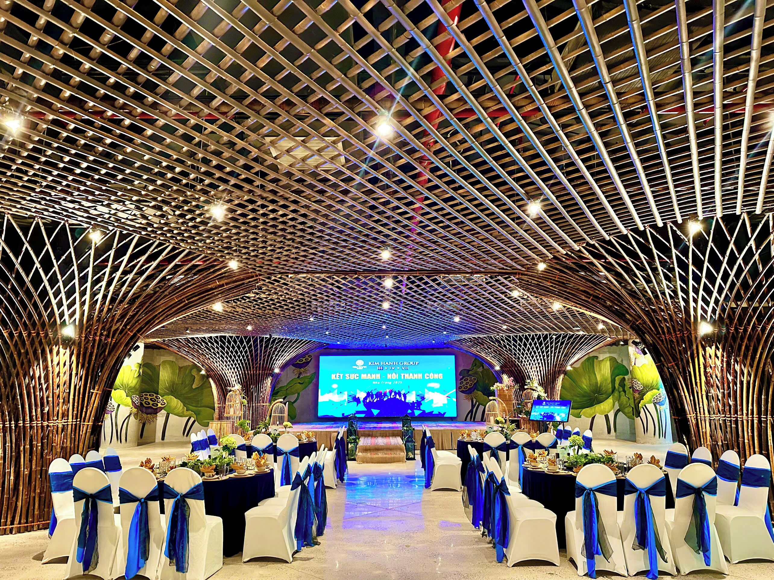 A banquet hall with a modern ceiling design, tables set for a formal event, and a stage with a large screen displaying text.