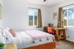 Une chambre avec un lit, un bureau et une chaise. Il y a des rideaux aux fenêtres et une plante sur le bureau.