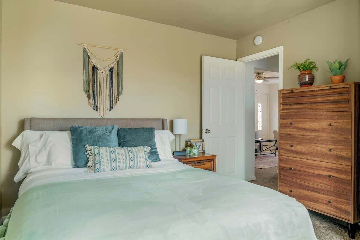 A bedroom with a bed, a wooden dresser, a macrame wall hanging, and a door leading to another room.
