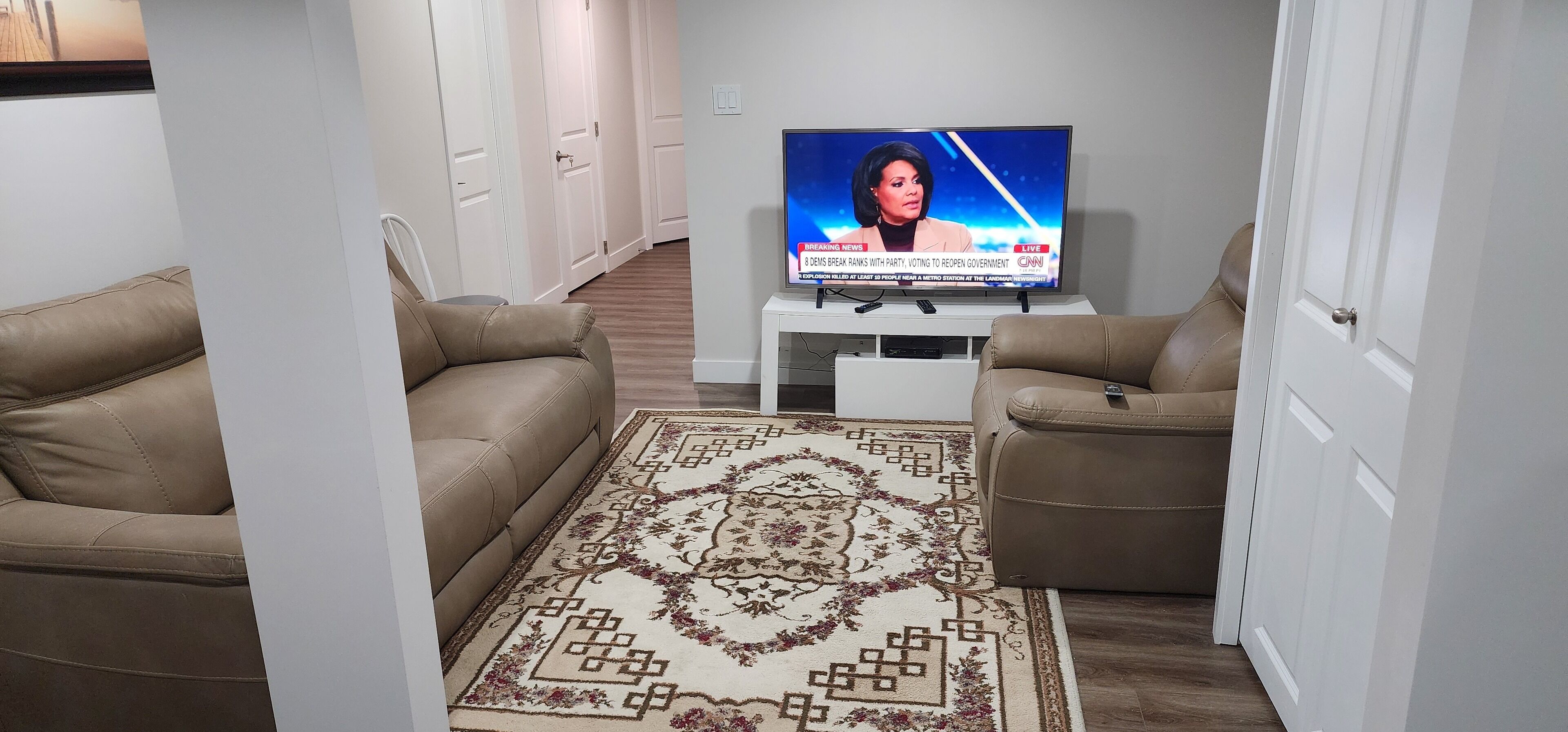 Ein Wohnzimmer mit zwei Ledersofas, einem gemusterten Teppich und einem Fernseher, der einen Nachrichtensender zeigt.