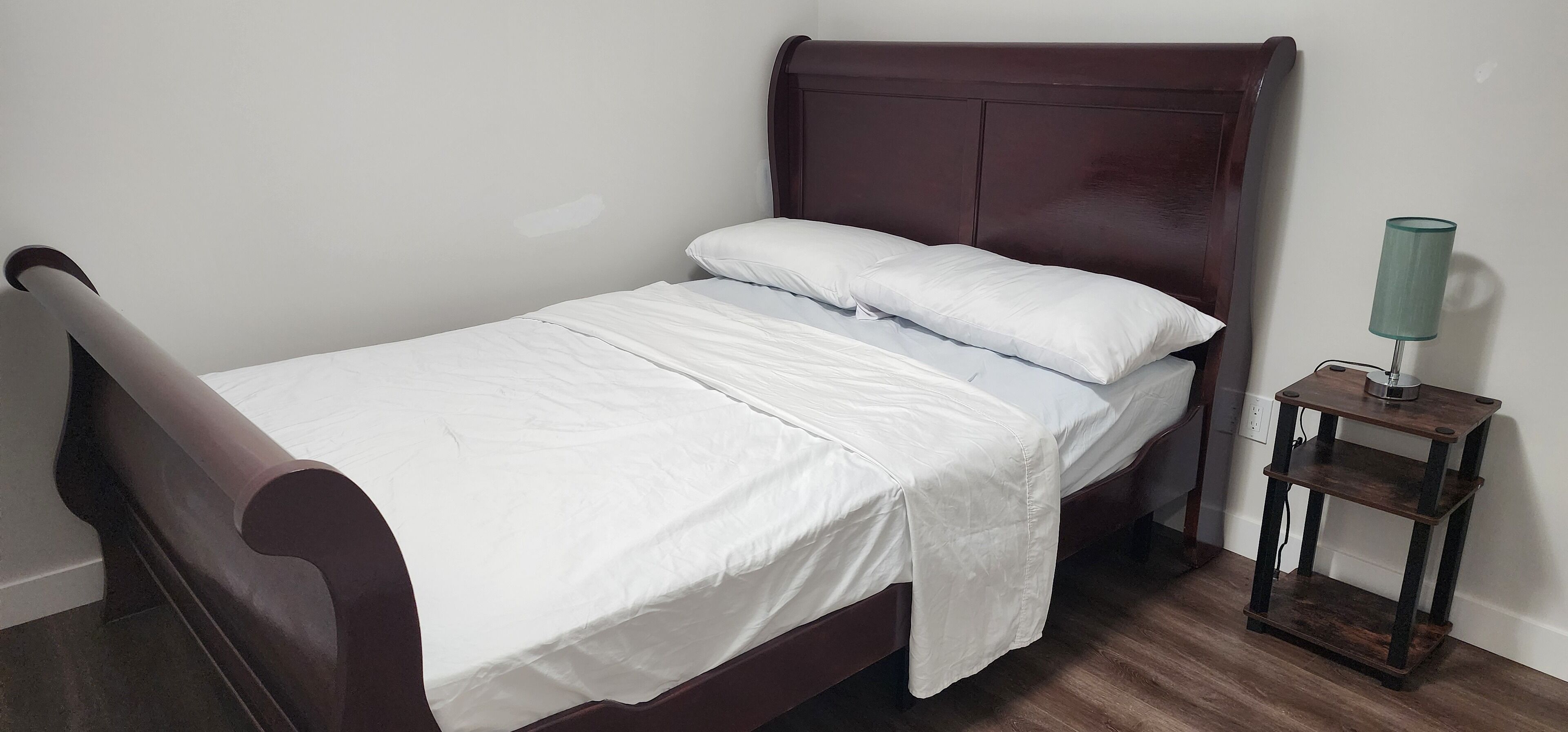 A single bed with a wooden headboard and white bedding, a bedside table with a lamp, and a wooden floor.