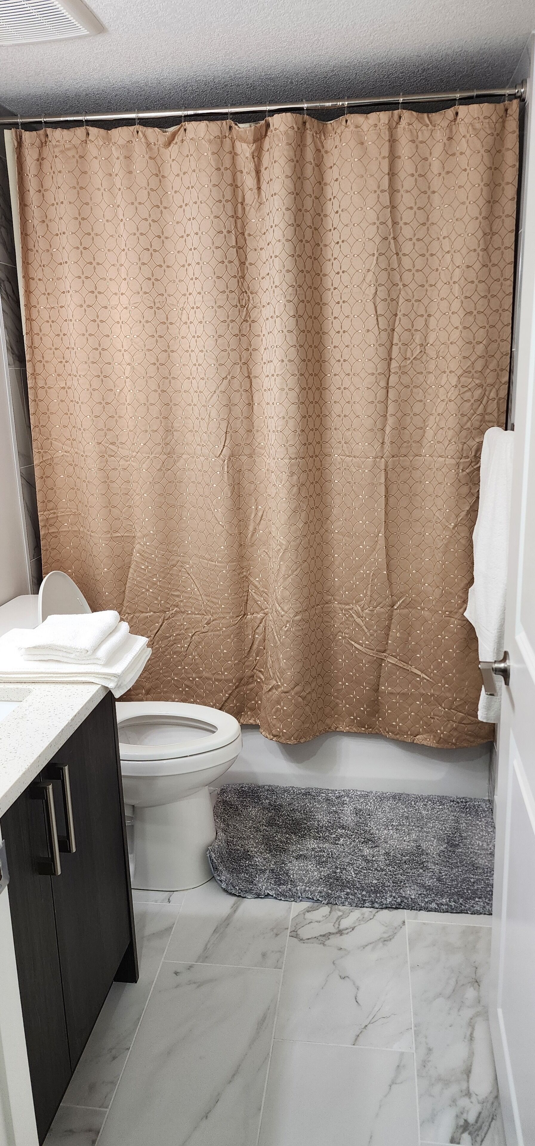 Un baño con inodoro, lavamanos con encimera blanca, armario oscuro y cortina de ducha estampada.