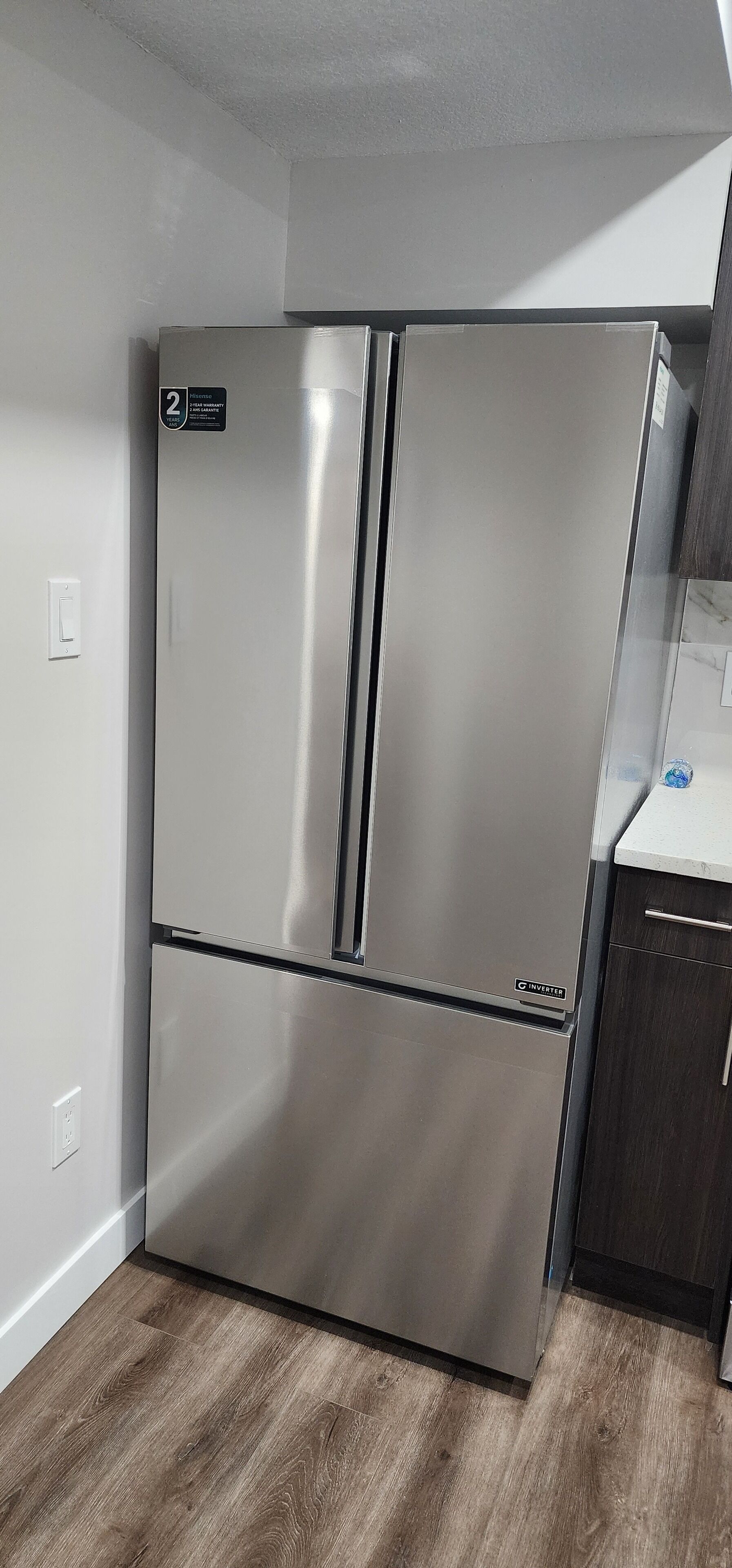 Un refrigerador de acero inoxidable en una cocina con piso de madera y paredes blancas.